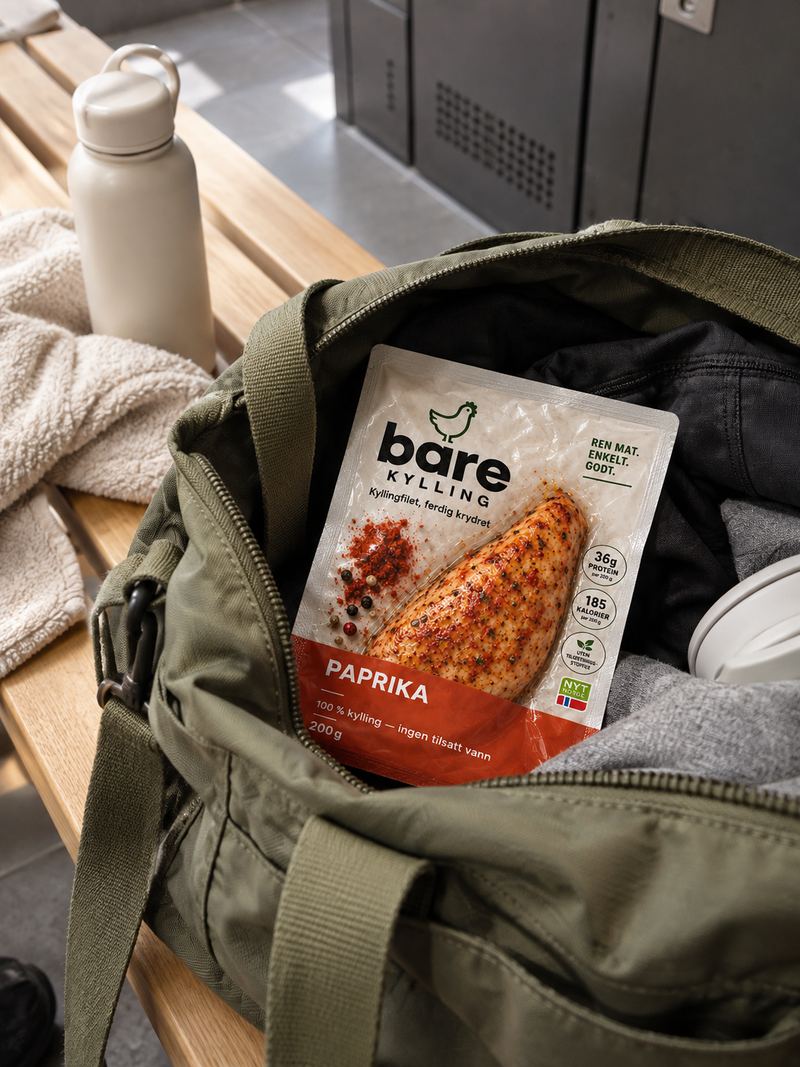 En pakke Bare Kylling Sweet Paprika oppi en åpen treningsbag på en benk i en garderobe, ved siden av en drikkeflaske og et håndkle.